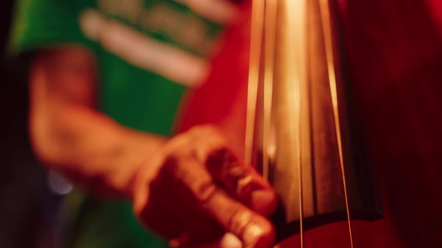 A musician's fingers glide along the strings of a double bass in slow motion, with the camera revealing his passionate performance under dramatic red stage lights