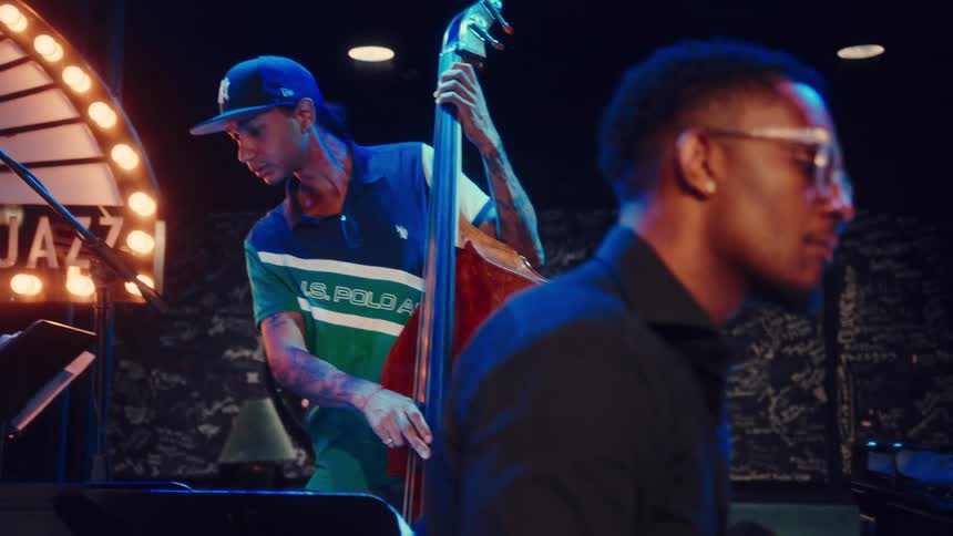 A tattooed bassist plays the double bass while a pianist performs alongside him during a live show in a moody jazz club