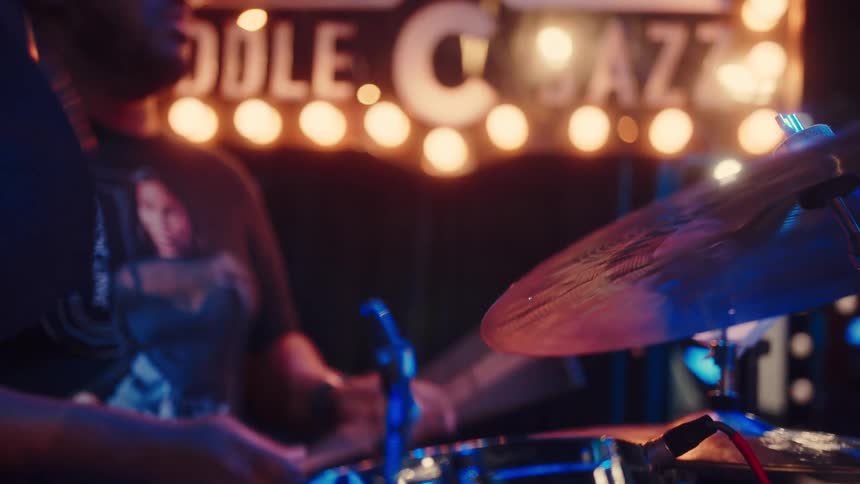 A jazz drummer plays passionately on a dimly lit stage, with the camera shifting focus to his cymbals