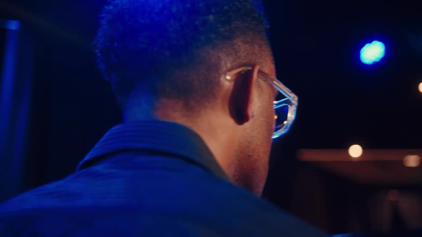 An over the shoulder close up of a musician with glasses playing in a dark club with moody blue lighting