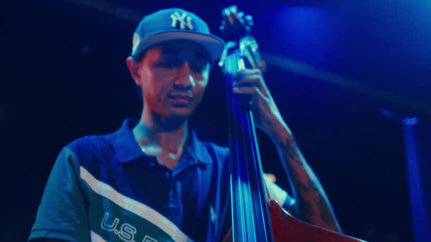 A tattooed musician in a baseball cap plays the double bass with intensity on a stage under cool blue and warm stage lights