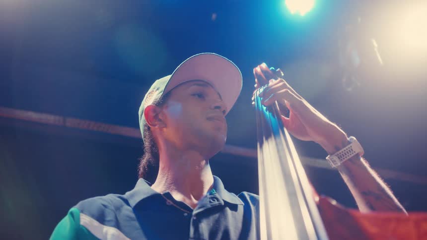 A young man with a backwards cap plays the upright bass with passion under colorful stage lights