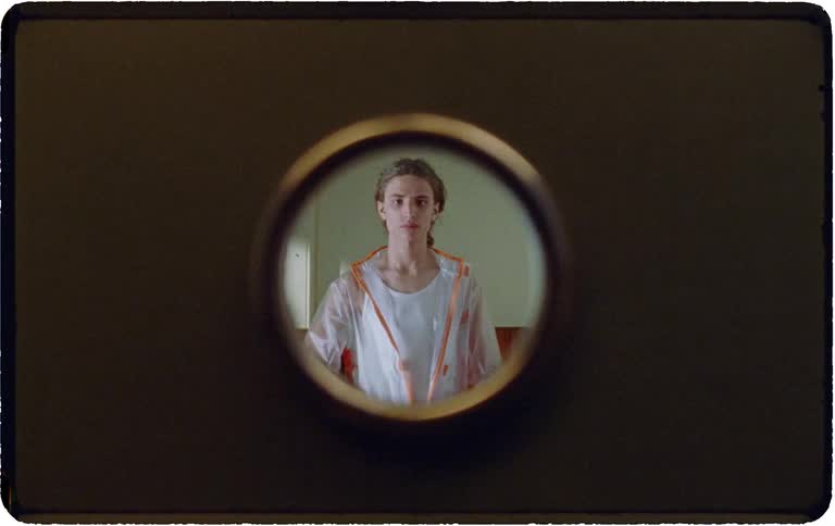 A young man in a transparent rain jacket approaches, stares into a door peephole, and then covers the lens