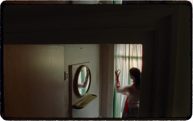 A woman seen from behind applies makeup while standing in a sunlit doorway at the end of a dark, vintage-style hallway