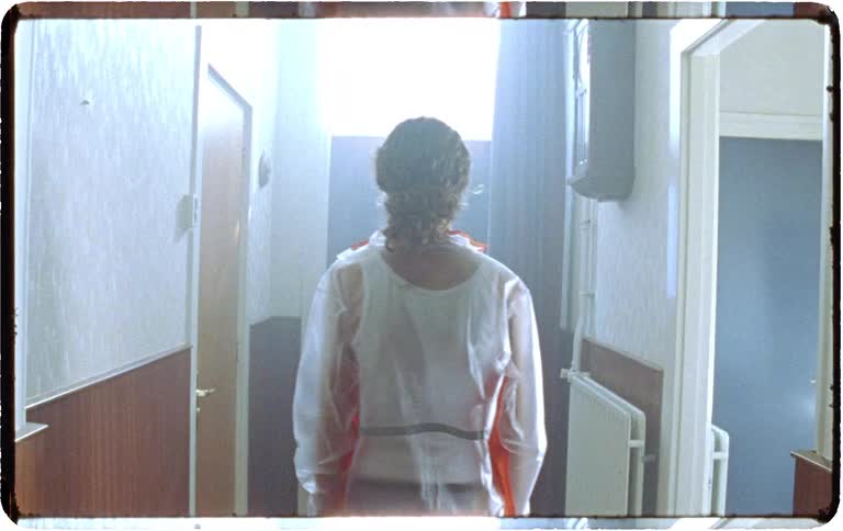 A woman wearing a transparent raincoat walks through a dark residential hallway towards a bright, mysterious light