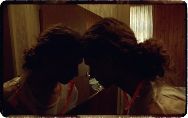 A silhouetted person in a dimly lit room leans forward to touch foreheads with their reflection in a mirror