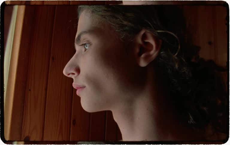 A pensive young man with long hair stands shirtless, thoughtfully gazing out of a window in a dimly lit, wood-paneled room