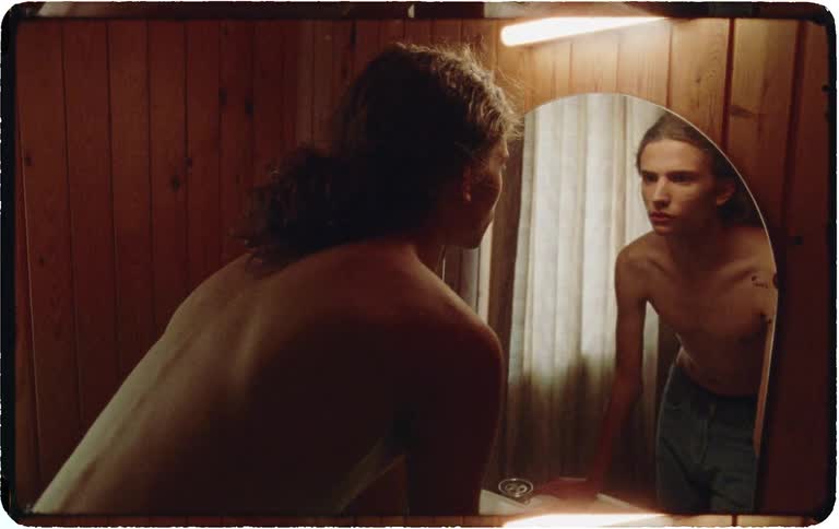 Over-the-shoulder shot of a shirtless young man with long hair looking at his reflection in a mirror in a dimly lit, vintage-style room