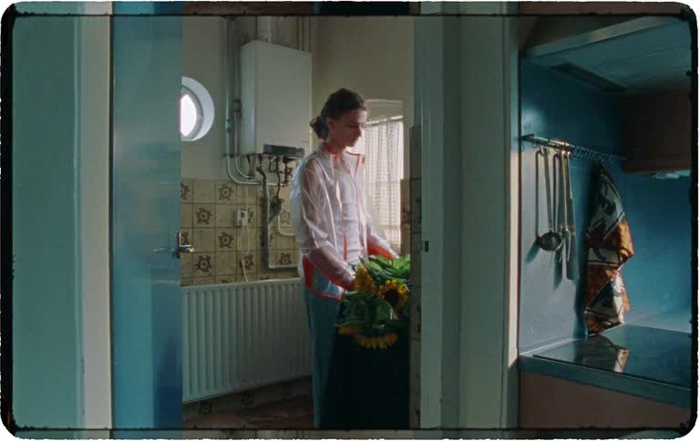 A pensive person in a translucent raincoat stands in a vintage kitchen holding a bouquet of sunflowers