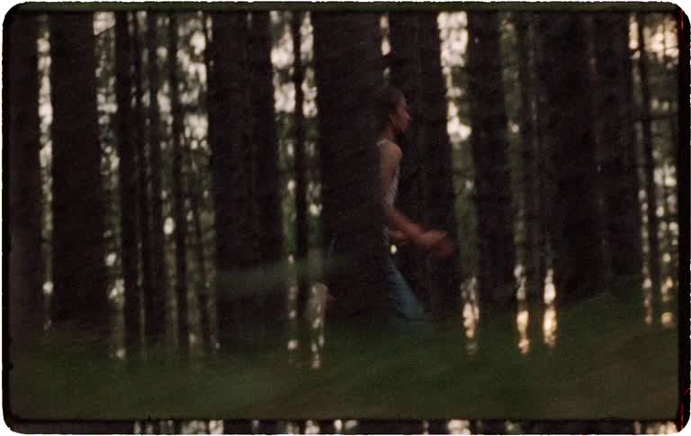 A grainy, vintage-style tracking shot follows a woman as she runs urgently through a dense forest at dusk