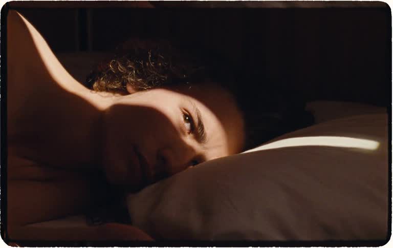 A close-up shot of a person with curly hair waking up slowly in bed as a ray of morning sunlight illuminates their face