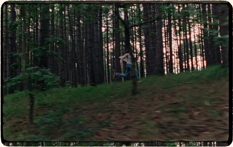 A person wearing jeans and suspenders runs frantically through a dense, dark forest as the camera tracks their movement in a vintage-style shot