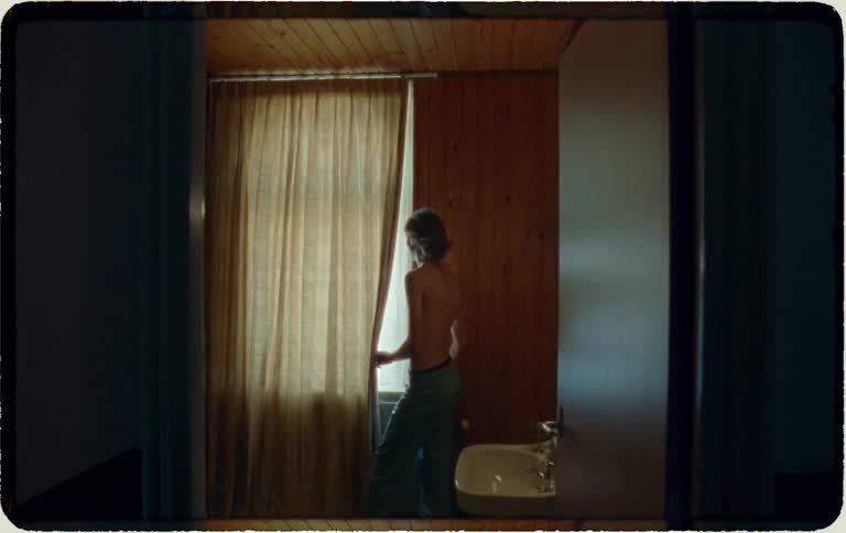 A woman stands in a dimly lit, vintage-style room, pulling back the curtain to peer outside