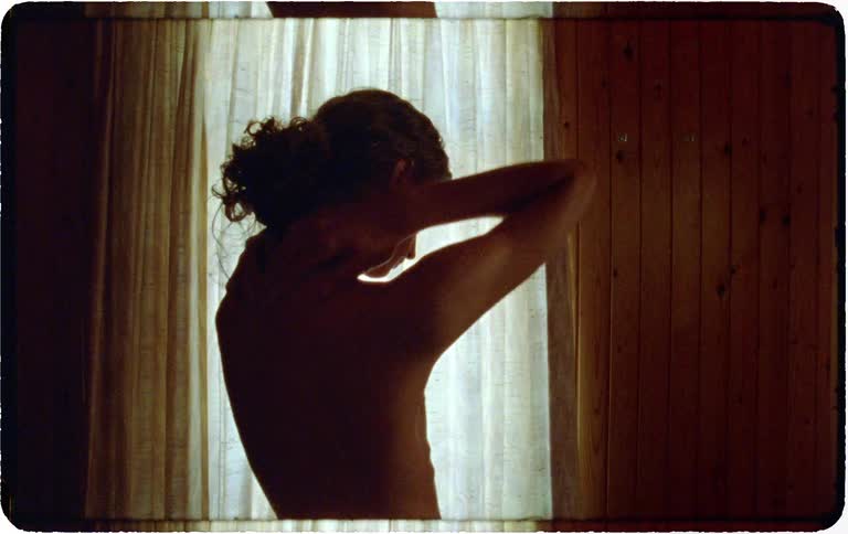 An introspective young person, first seen in a mirror, stands shirtless in silhouette by a sunlit window in a rustic, wood-paneled room