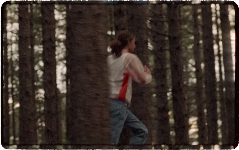 A vintage-style tracking shot follows a man with long hair as he runs through a dense forest at dusk