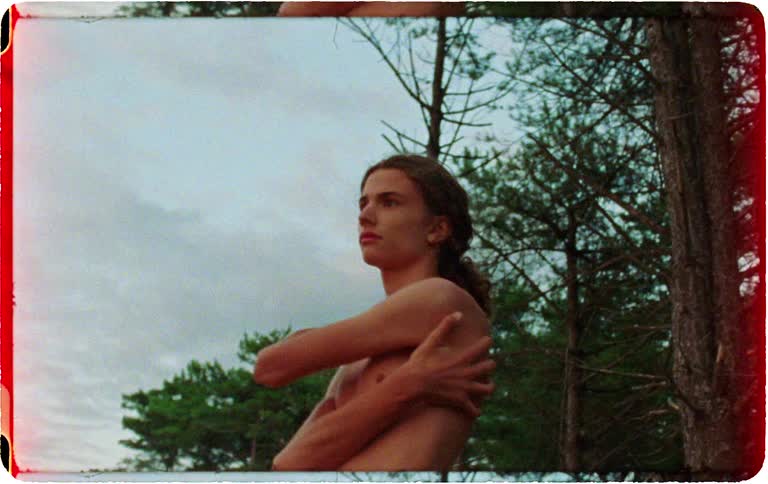 A pensive, shirtless young man with long hair stands alone in nature against a cloudy sky in a grainy, vintage-style film