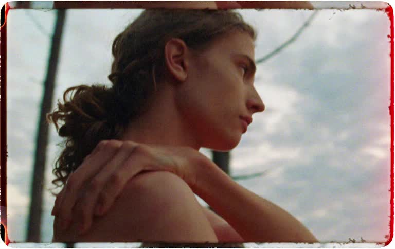 Vintage-style, low-angle shot of a contemplative young person standing shirtless amongst trees under a cloudy sky
