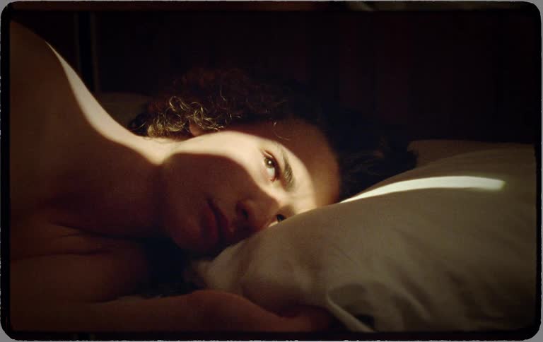 A person with curly hair slowly wakes up in bed as a sliver of morning sunlight shines across their face
