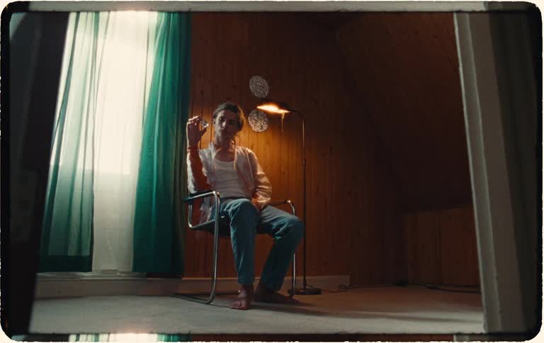 A thoughtful young man sits alone in a vintage-style attic room, holding a cigarette and looking at the camera