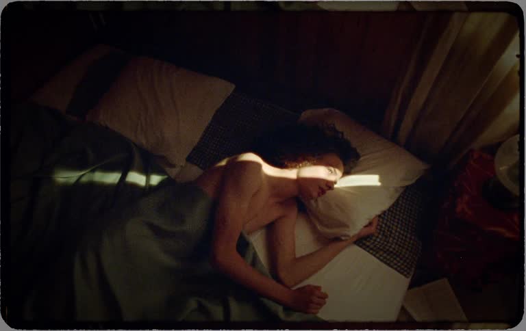 A vintage-style time-lapse of a young man waking up in bed as a beam of morning sun moves across his body