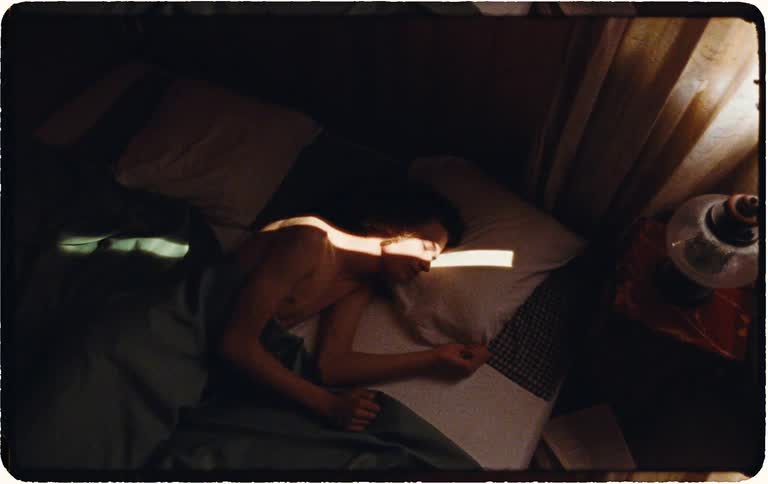 High angle shot of a man sleeping in a dark bedroom as a beam of sunlight moves across him, causing him to stir and turn over