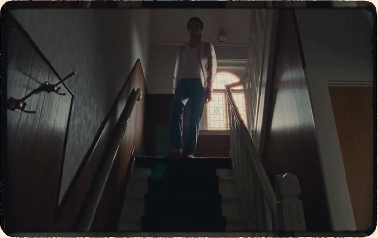 A young person slowly walks down the stairs in a moody, backlit hallway in this vintage-style footage