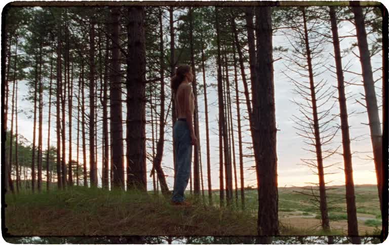 Vintage-style footage shows a young man walking up a hill in a pine forest, taking off his shirt and standing pensively at dusk