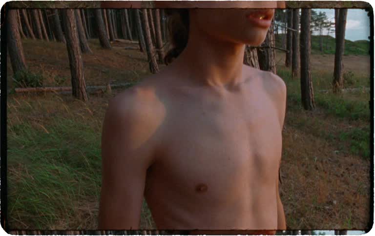Young man stands topless among coastal pine trees at sunset gazing over dunes and sea