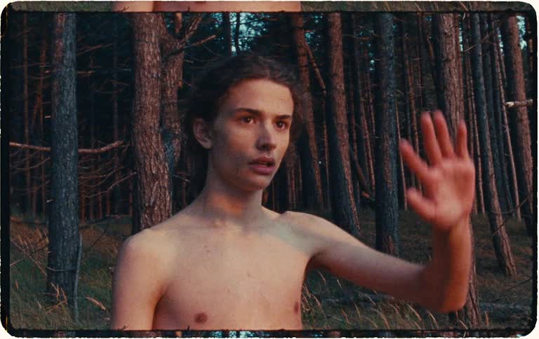 Shirtless young man walks through a pine forest at dusk and raises his hand in a moody vintage close-up