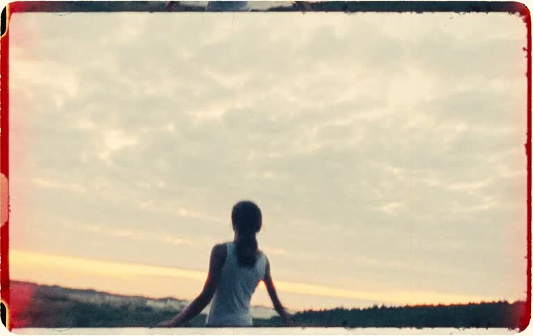 A young man with long hair runs frantically out of a dark forest into an open field at sunset, captured on vintage film