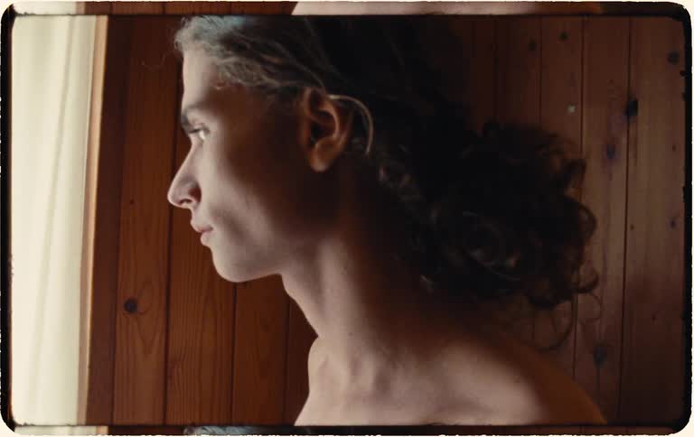 Slow zoom-in on a pensive young man standing shirtless by a window in a rustic, wood-paneled room, bathed in natural light