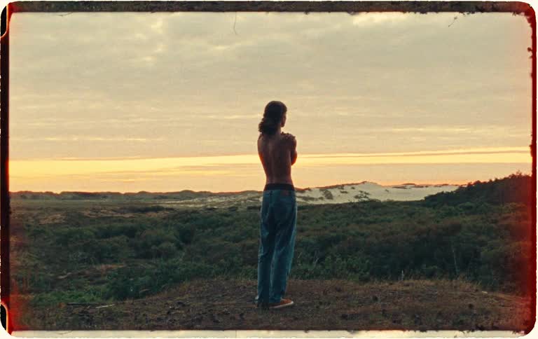 A lone, shirtless figure contemplates the vast coastal landscape at sunset in this vintage-style film clip