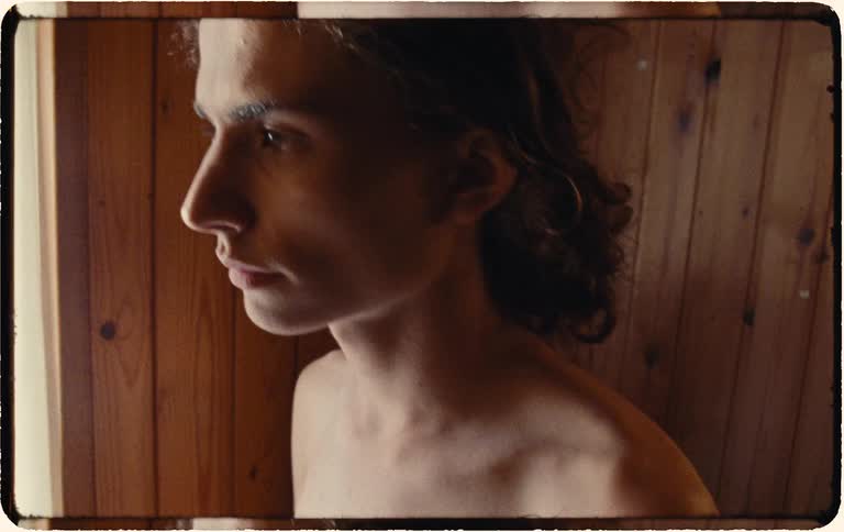 A slow zoom out from a pensive young man with long hair looking out a window in a rustic, wood-paneled room