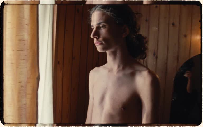 Young person stands shirtless by a sunlit window in a wood-paneled room with a contemplative mood