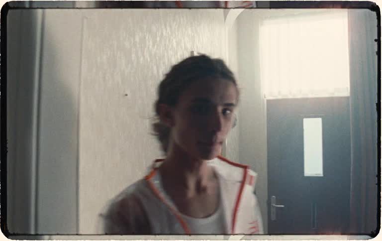 Moody, grainy super 8 footage of a pensive young person with braided hair standing in a softly lit hallway