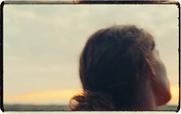 An intimate, vintage-style close-up of a person gently caressing their shoulder and neck while looking out at a serene landscape during sunset