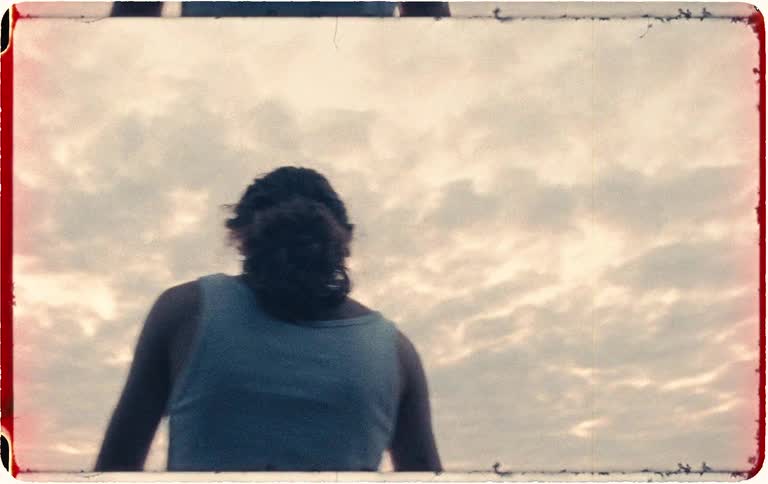Dreamy vintage-style footage shows a young person with long hair walking and stretching their arms towards a vast, cloudy sky at sunset