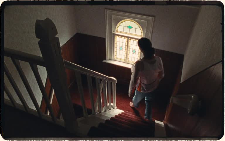 High-angle vintage shot of a woman slowly carrying her baby down a dimly lit staircase towards a stained-glass window