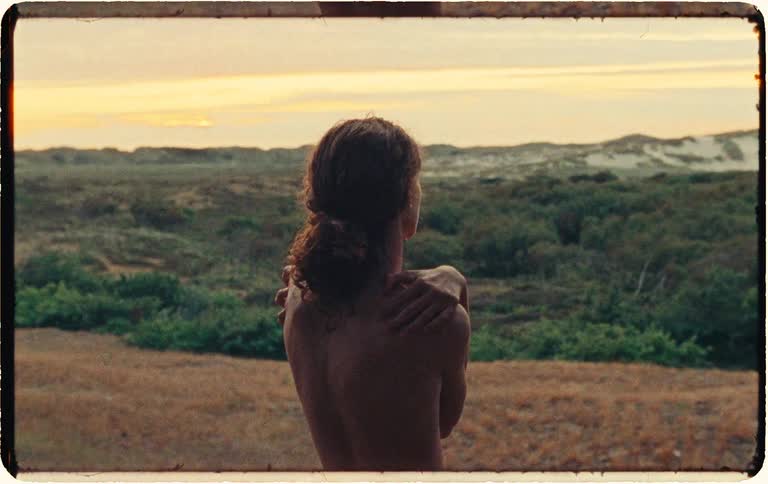 Vintage film footage shows a nude woman with her arms crossed, gazing at a beautiful sunset over a serene natural landscape