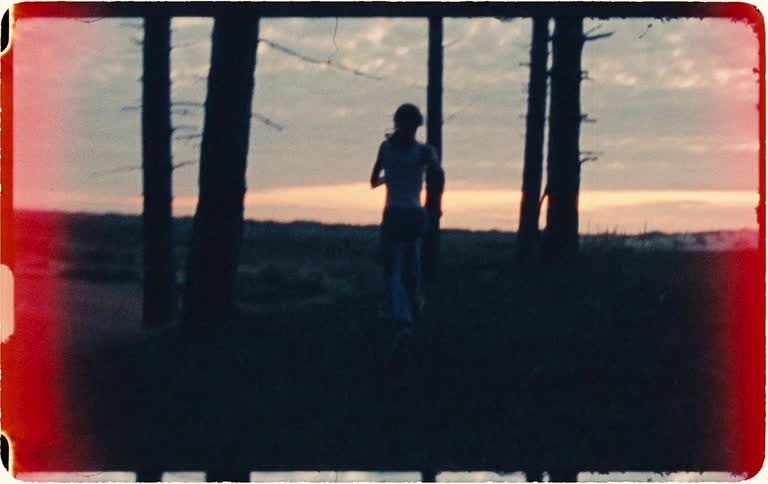 Vintage 8mm film captures a silhouetted figure running away through a sparse coastal forest at sunset, creating a moody and nostalgic scene