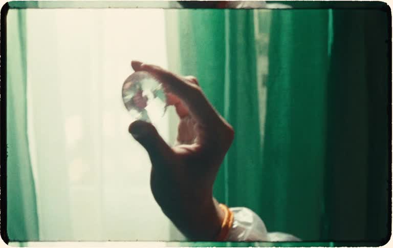 A hand holds up a small, faceted crystal to the light, creating shimmering reflections in a retro-style, grainy film aesthetic