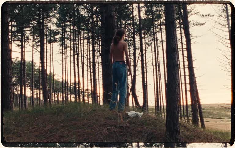 Vintage film shows a young man with long hair walking through a pine forest, stopping on a hill to take off his shirt and enjoy the sunset