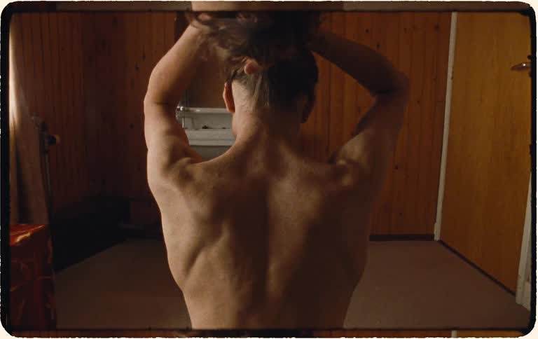 Rear view of a shirtless person with long curly hair styling it into a bun in a rustic room
