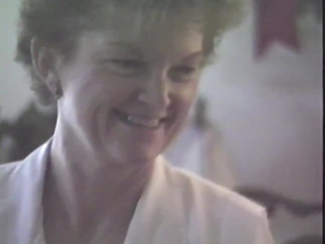 A warm and friendly older woman with short hair smiles and talks to the camera in a vintage home video
