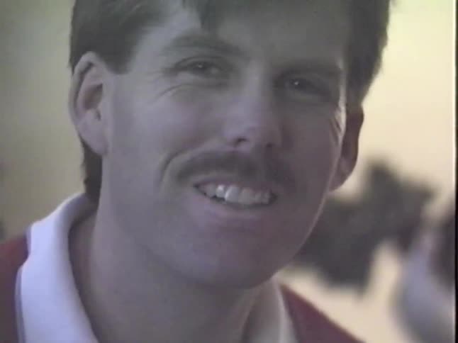 A close-up of a man with a mustache making a silly face and smiling at the camera in a grainy home video