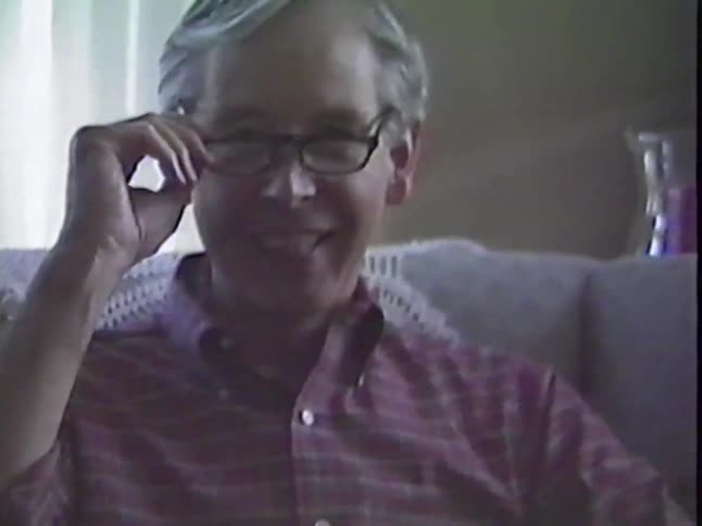 A smiling older man with gray hair takes off his glasses while sitting on a couch in a warm, archival home video