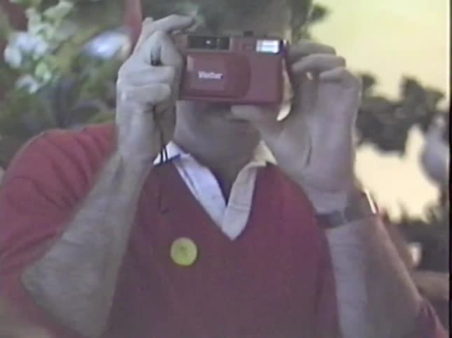 A man with a mustache wearing a red sweater takes a picture with his new retro red Vivitar camera during a holiday gathering
