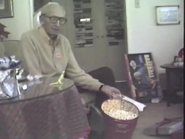 An elderly grandfather smiles as he opens a large tin of popcorn for Christmas in a vintage home movie