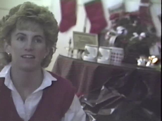 A woman in a red sweater vest opens a Christmas present and reacts with joy and surprise, from a vintage home video