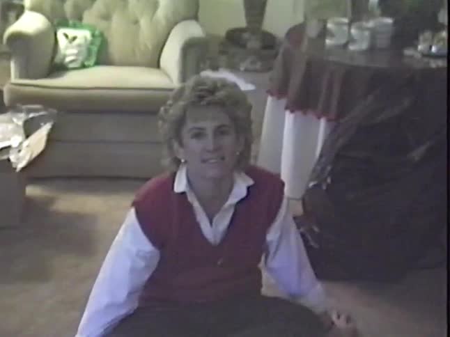 A woman with a classic 80s hairstyle smiles and laughs while sitting on the floor of a living room decorated for Christmas in this grainy vintage home video